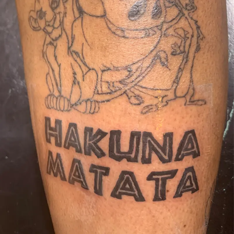 Bold block lettering tattoo reading "Hakuna Matata" below a Lion King outline on the leg, by Andres at Killer Silver Tattoo