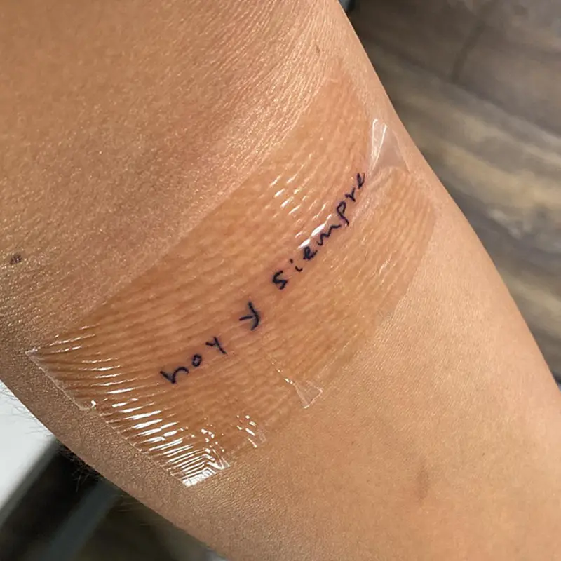 Fine-line handwritten tattoo reading "hoy y siempre" on the arm under a clear wound cover, by Andres at Killer Silver Tattoo