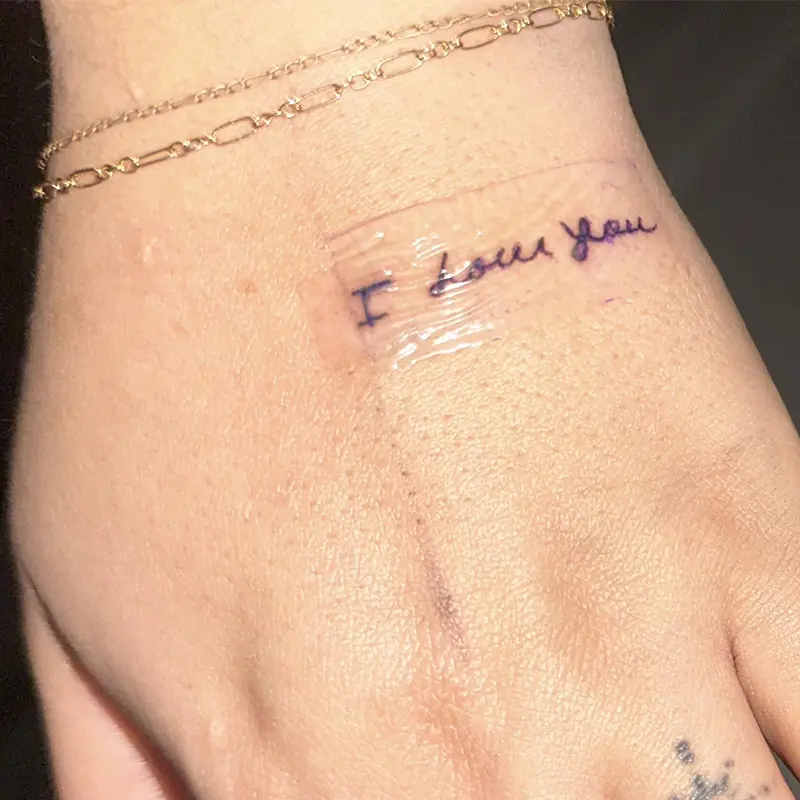 Fine-line handwritten tattoo reading "I love you" on the back of the hand, by Andres at Killer Silver Tattoo