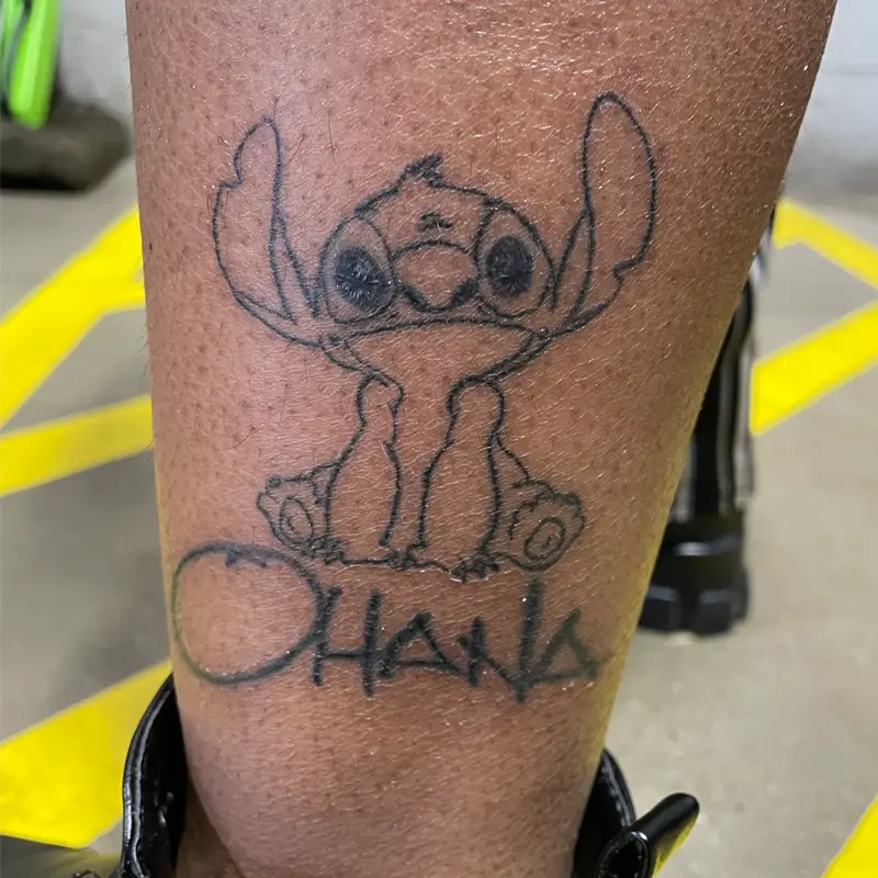 Fine-line outline tattoo of Stitch above "Ohana" lettering on the lower leg, by Andres at Killer Silver Tattoo