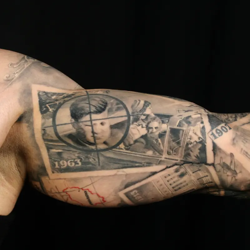 Black and gray realism tattoo of the 1963 Kennedy motorcade under a rifle crosshair, collaged with a map and newspaper on the upper arm