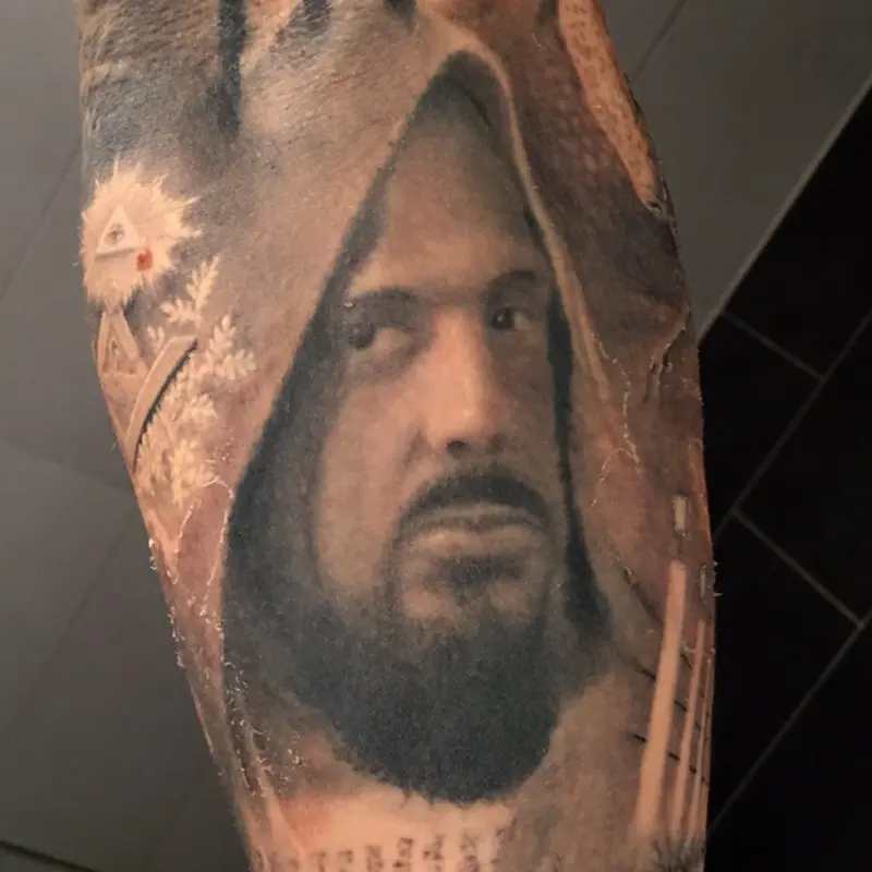 Black and gray realism portrait tattoo of a hooded, bearded Judas Iscariot on the forearm, with the Eye of Providence at left