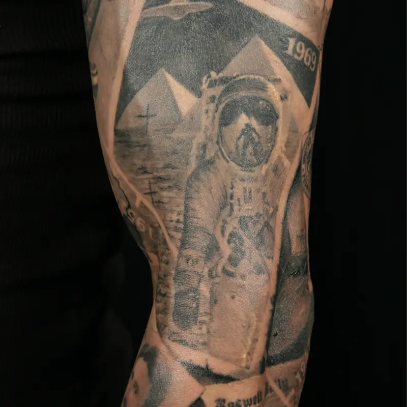 Black and gray realism tattoo of an astronaut on the moon, pyramids behind him and an alien reflected in his visor, dated 1969