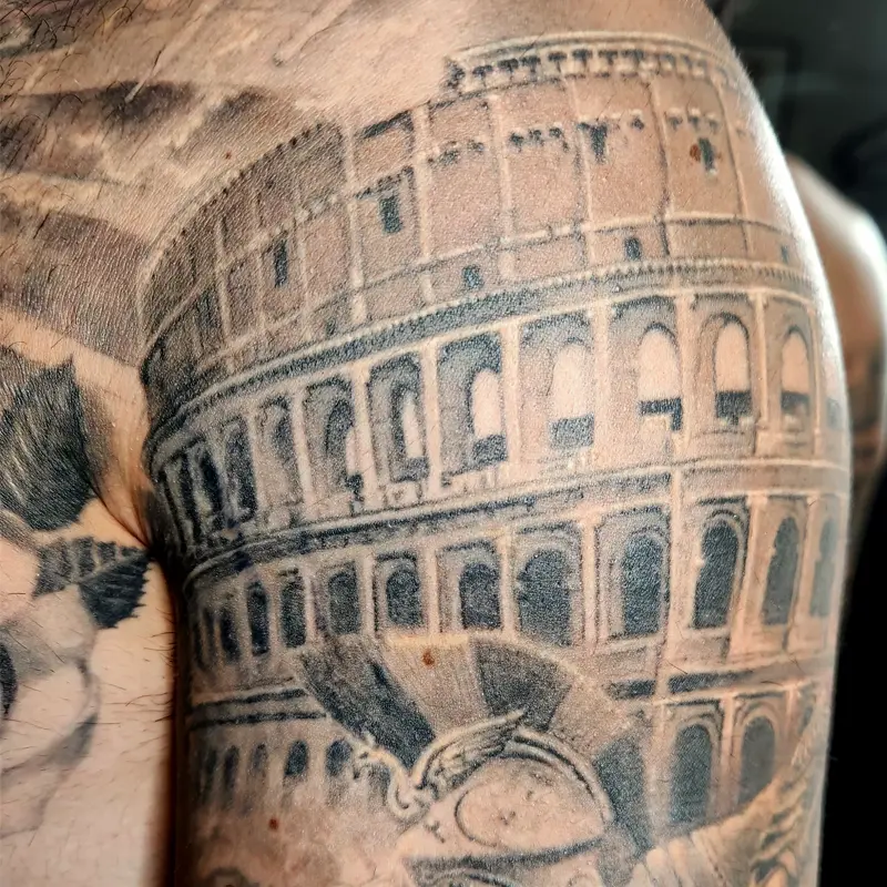 Black and gray realism tattoo of the Roman Colosseum across the shoulder, above a crested Roman helmet with a winged emblem