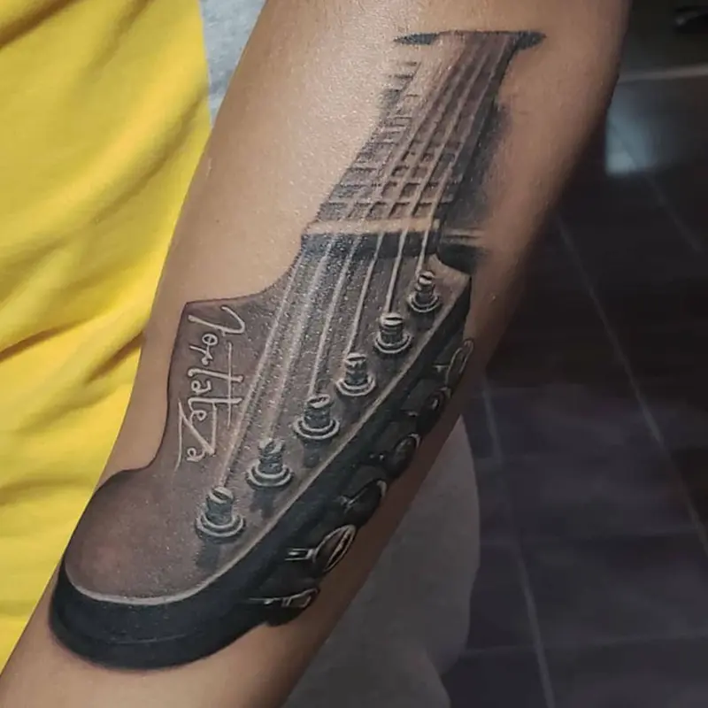 Black and gray realism tattoo of a guitar headstock with the name Fortaleza, by Philipus